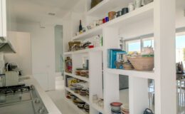 Modern kitchen interior featuring a white shelving unit filled with colorful plates, mugs, and glassware, alongside neatly stacked cookbooks and decorative items. The minimalist design enhances the spacious feel of the kitchen, with natural light streaming through large windows.