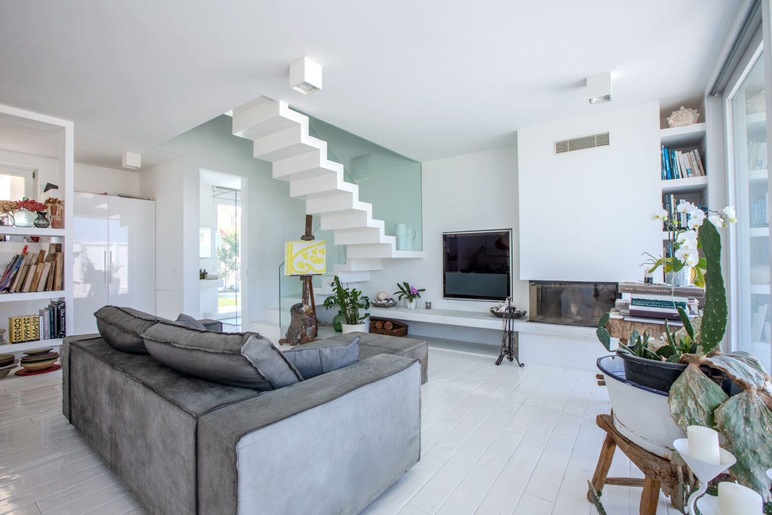 Modern living room featuring a grey sofa, minimalist decor, and a stylish staircase. The space includes a flat-screen TV, a decorative fireplace, and plants, creating a bright and inviting atmosphere. | Only on SopranoVillas