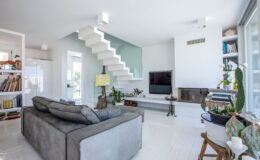 Modern living room featuring a grey sofa, minimalist decor, and a stylish staircase. The space includes a flat-screen TV, a decorative fireplace, and plants, creating a bright and inviting atmosphere.