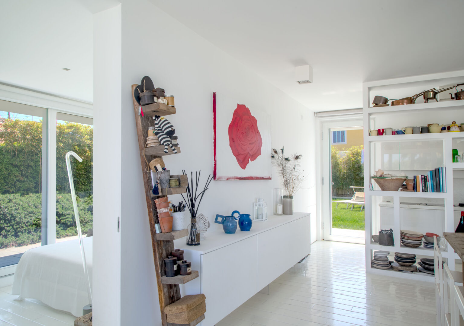 Modern interior design featuring a bright, minimalist space with white walls and polished floors. A wooden shelf displays various decorative items and plants, while a large red rose painting adds a pop of color. The room has large windows that provide natural light and a view of the outdoor greenery. | Only on SopranoVillas