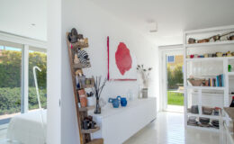 Modern interior design featuring a bright, minimalist space with white walls and polished floors. A wooden shelf displays various decorative items and plants, while a large red rose painting adds a pop of color. The room has large windows that provide natural light and a view of the outdoor greenery.