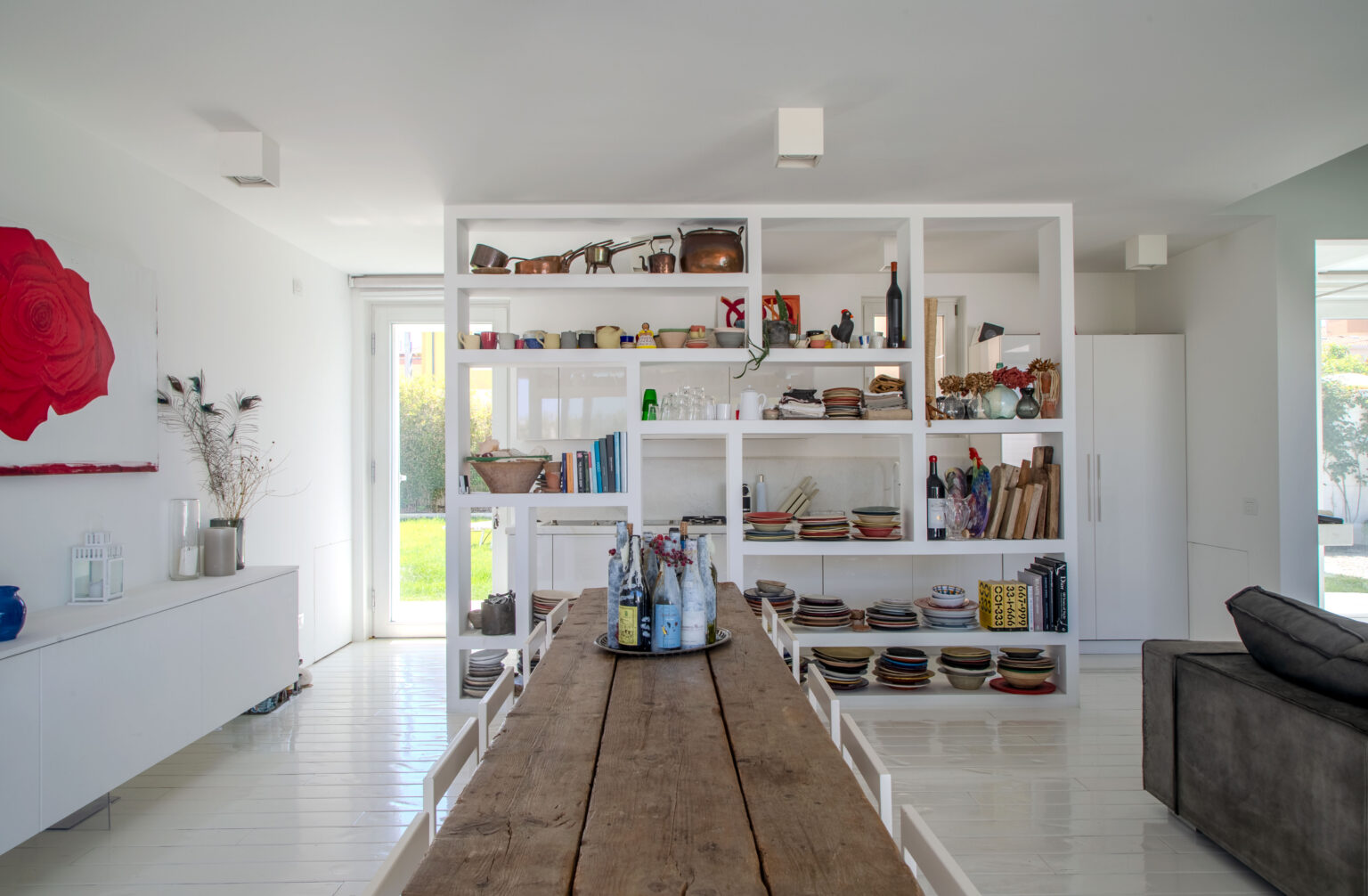 Modern kitchen and dining area featuring a rustic wooden table, white shelving filled with colorful dishes and kitchenware, and a large window allowing natural light. The decor includes a red rose artwork and a minimalist aesthetic, creating a bright and inviting space. | Only on SopranoVillas