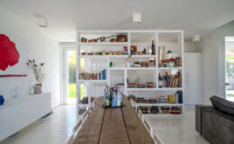 Modern kitchen and dining area featuring a rustic wooden table, white shelving filled with colorful dishes and kitchenware, and a large window allowing natural light. The decor includes a red rose artwork and a minimalist aesthetic, creating a bright and inviting space.