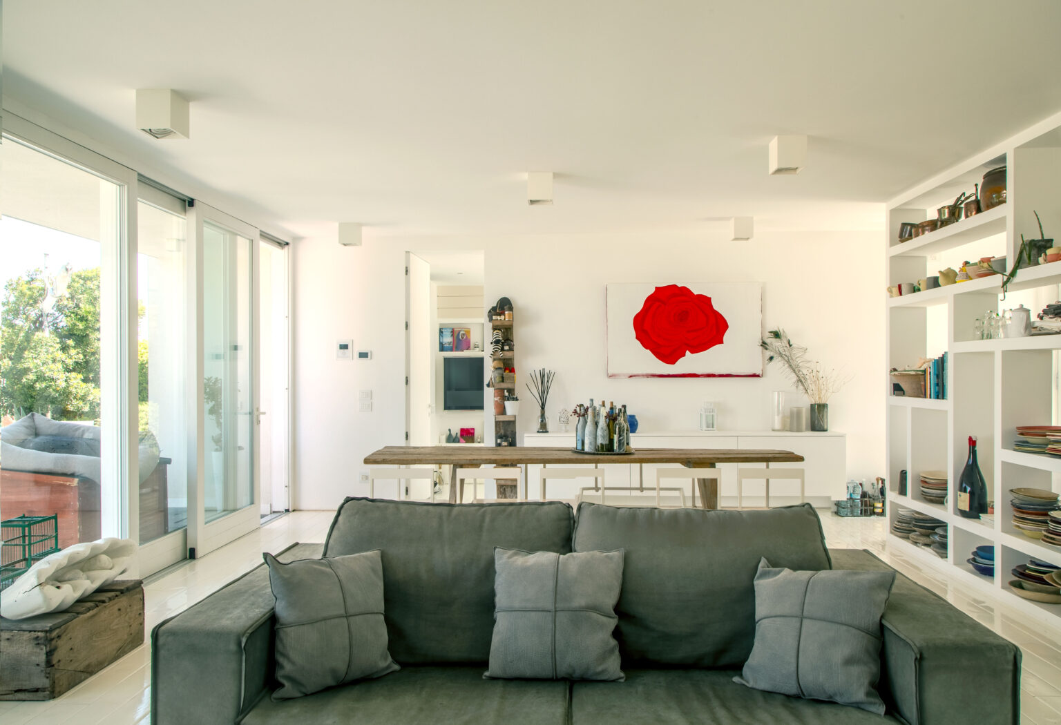 Modern living room featuring a grey sofa with decorative pillows, a large red rose painting on the wall, and a wooden dining table. The space includes glass doors leading to an outdoor area, built-in shelves displaying various items, and a bright, airy atmosphere. | Only on SopranoVillas