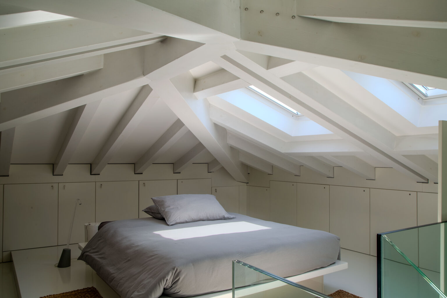Bright and modern attic bedroom featuring a spacious bed with gray bedding, surrounded by minimalist storage cabinets and illuminated by skylights, showcasing clean lines and a contemporary design. | Only on SopranoVillas