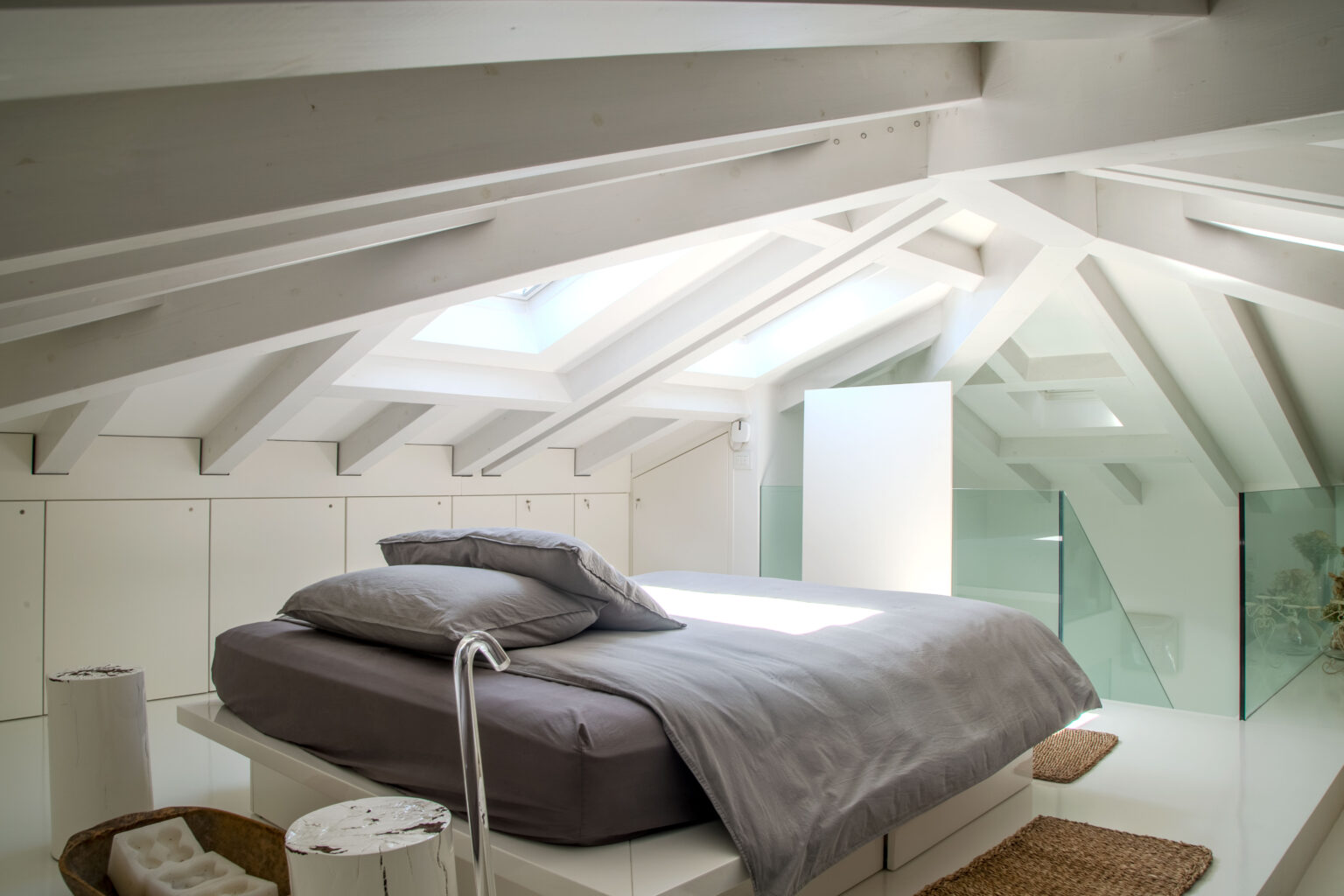 Modern attic bedroom featuring a minimalist design, white wooden beams, a large skylight, and a neatly made bed with gray bedding. The space includes glass accents and natural light, creating a bright and airy atmosphere. | Only on SopranoVillas