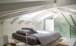 Modern attic bedroom featuring a minimalist design, white wooden beams, a large skylight, and a neatly made bed with gray bedding. The space includes glass accents and natural light, creating a bright and airy atmosphere.
