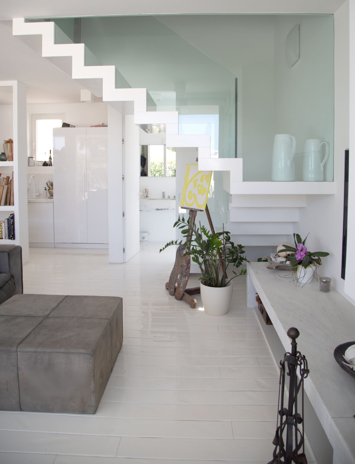 Modern interior design featuring a white staircase with glass railing, a gray ottoman, and a potted plant. The open layout includes a kitchen area with white cabinetry, light hardwood floors, and minimalist decor elements. | Only on SopranoVillas