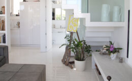 Modern interior design featuring a white staircase with glass railing, a gray ottoman, and a potted plant. The open layout includes a kitchen area with white cabinetry, light hardwood floors, and minimalist decor elements.