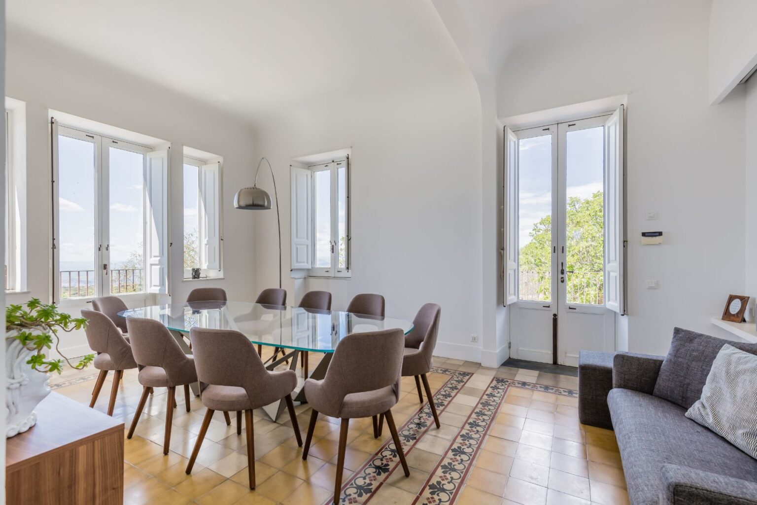 Modern dining room featuring a glass table surrounded by eight stylish chairs, large windows providing natural light, and a cozy sofa in a well-decorated space with elegant tile flooring and greenery. Ideal for gatherings and meetings. | Only on SopranoVillas
