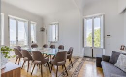 Modern dining room featuring a glass table surrounded by eight stylish chairs, large windows providing natural light, and a cozy sofa in a well-decorated space with elegant tile flooring and greenery. Ideal for gatherings and meetings.