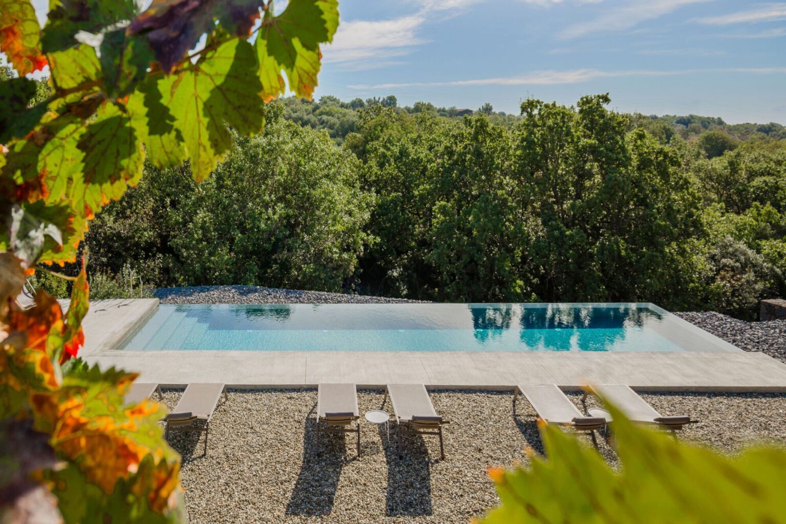 Scenic view of a modern infinity pool surrounded by lush greenery, featuring lounge chairs on a gravel patio, ideal for relaxation and outdoor enjoyment. | Only on SopranoVillas