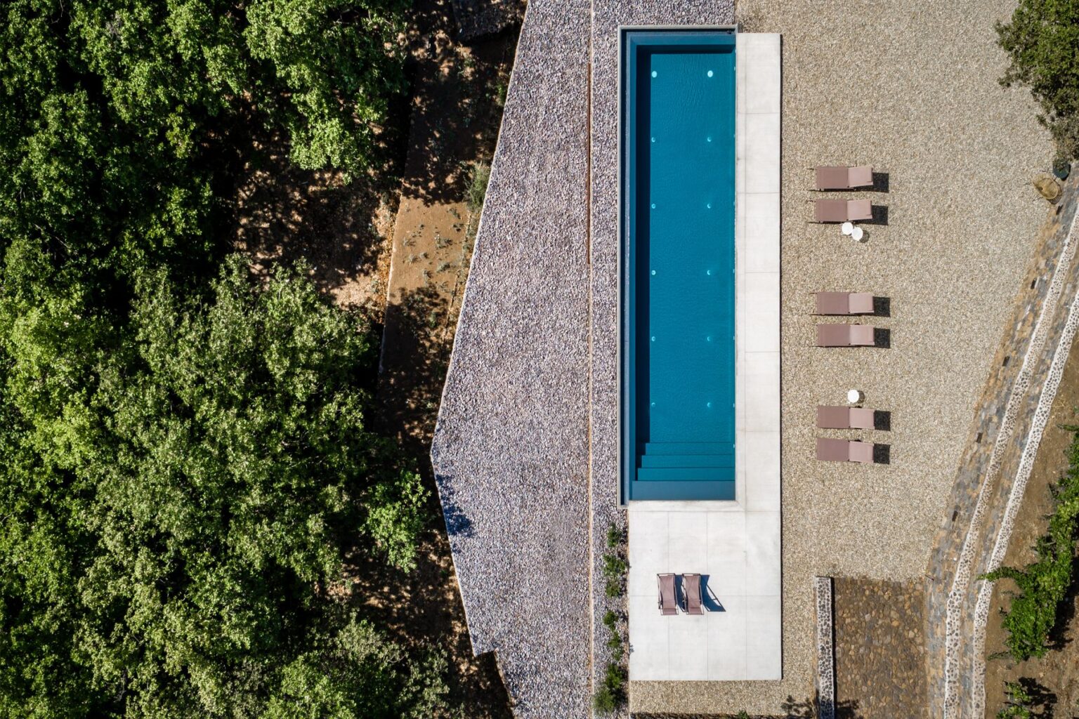 Aerial view of a modern swimming pool surrounded by a landscaped area featuring gravel and lounge chairs, set amidst lush green trees. The pool's clean lines and contemporary design create a serene outdoor space ideal for relaxation and leisure. | Only on SopranoVillas