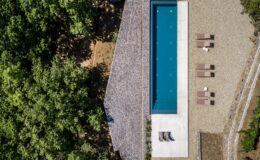 Aerial view of a modern swimming pool surrounded by a landscaped area featuring gravel and lounge chairs, set amidst lush green trees. The pool's clean lines and contemporary design create a serene outdoor space ideal for relaxation and leisure.