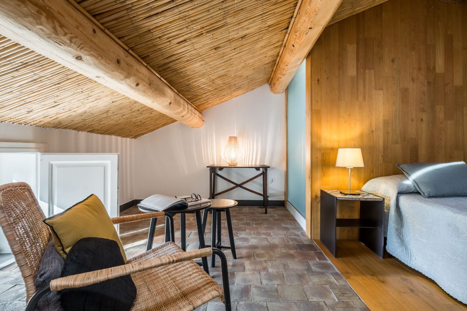 Cozy attic bedroom featuring a sloped bamboo ceiling, a comfortable bed with soft linens, a stylish bedside table with a lamp, and a reading nook with a woven chair and a side table. Natural light enhances the warm wooden accents and rustic floor tiles, creating a serene atmosphere. | Only on SopranoVillas