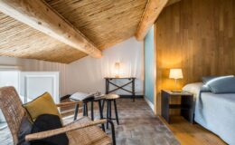 Cozy attic bedroom featuring a sloped bamboo ceiling, a comfortable bed with soft linens, a stylish bedside table with a lamp, and a reading nook with a woven chair and a side table. Natural light enhances the warm wooden accents and rustic floor tiles, creating a serene atmosphere.