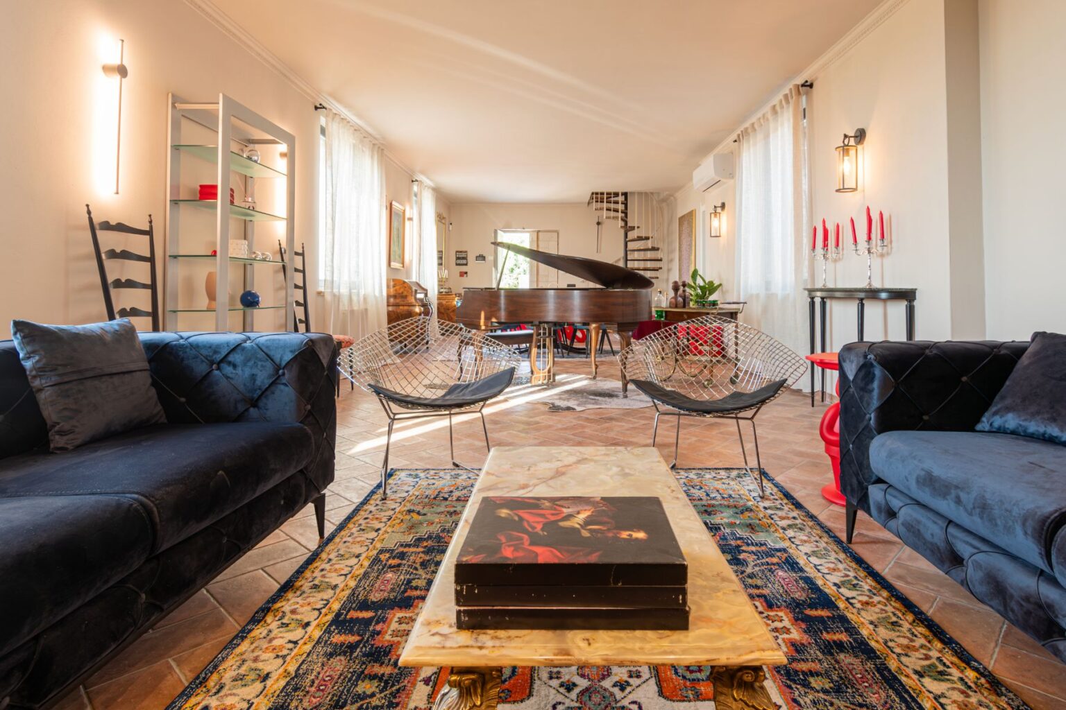 Spacious and stylish living room featuring a grand piano, plush velvet sofas, and contemporary wire chairs. The room is illuminated by natural light through sheer curtains, highlighting a decorative coffee table and an elegant rug. A modern shelving unit displays decorative items, while red candles add a pop of color to the decor. | Only on SopranoVillas