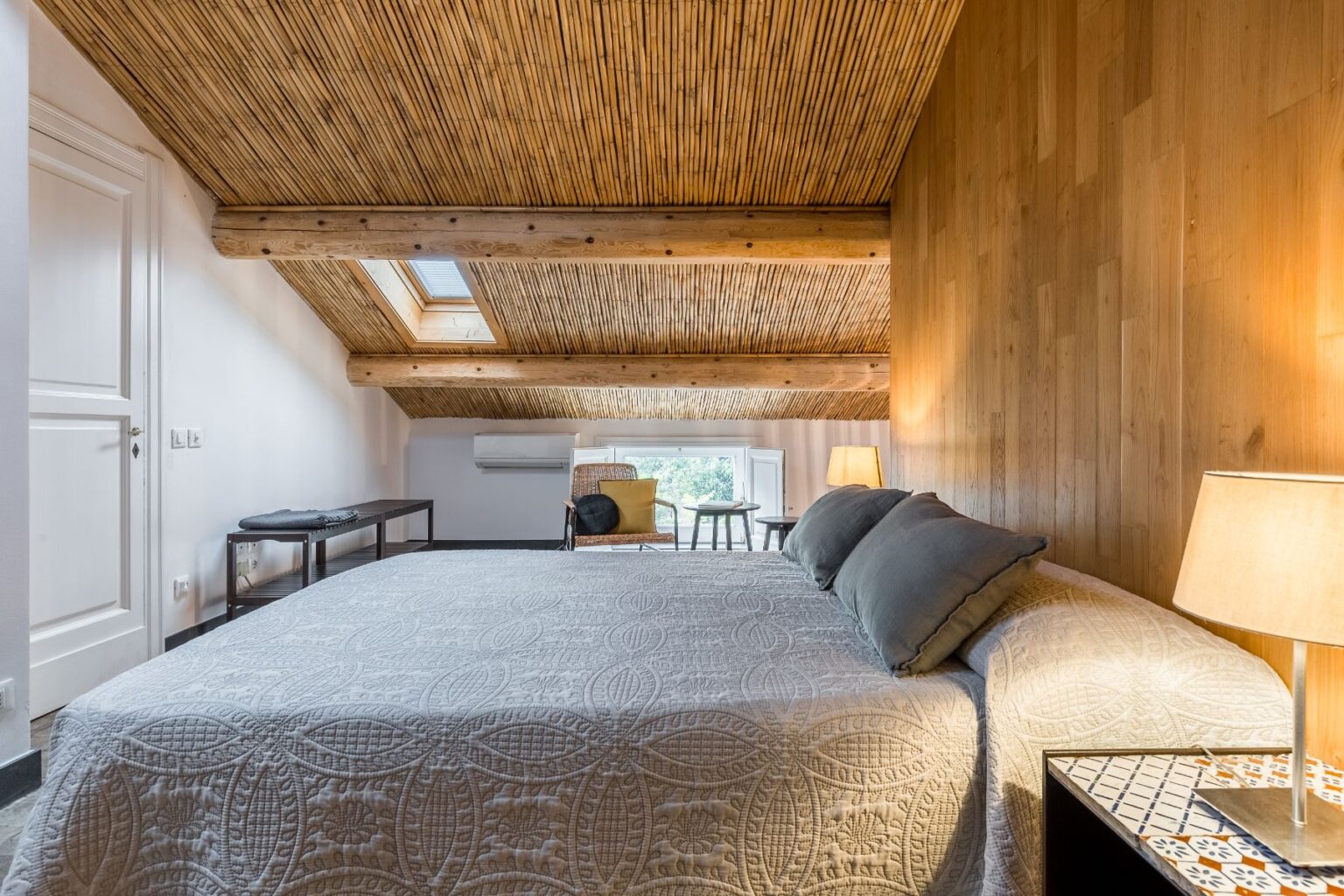 Cozy attic bedroom featuring a large bed with decorative quilt, wooden walls, and a bamboo ceiling. Natural light streams through a skylight, illuminating a seating area with a chair and side table. A bedside lamp adds warmth to the inviting atmosphere. | Only on SopranoVillas