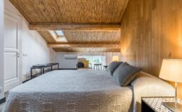 Cozy attic bedroom featuring a large bed with decorative quilt, wooden walls, and a bamboo ceiling. Natural light streams through a skylight, illuminating a seating area with a chair and side table. A bedside lamp adds warmth to the inviting atmosphere.