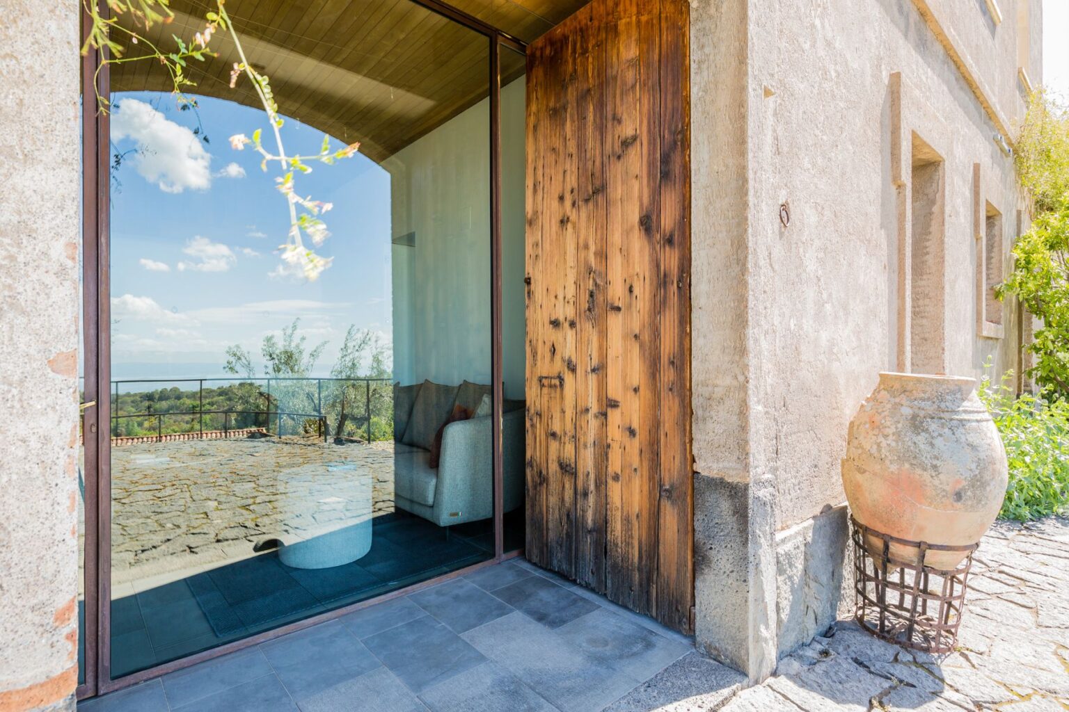 Modern entrance of a rustic house featuring a large glass door, wooden accents, and a view of the surrounding landscape. The stone patio and decorative pottery enhance the inviting atmosphere, perfect for showcasing outdoor living spaces. | Only on SopranoVillas