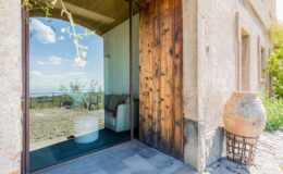Modern entrance of a rustic house featuring a large glass door, wooden accents, and a view of the surrounding landscape. The stone patio and decorative pottery enhance the inviting atmosphere, perfect for showcasing outdoor living spaces.