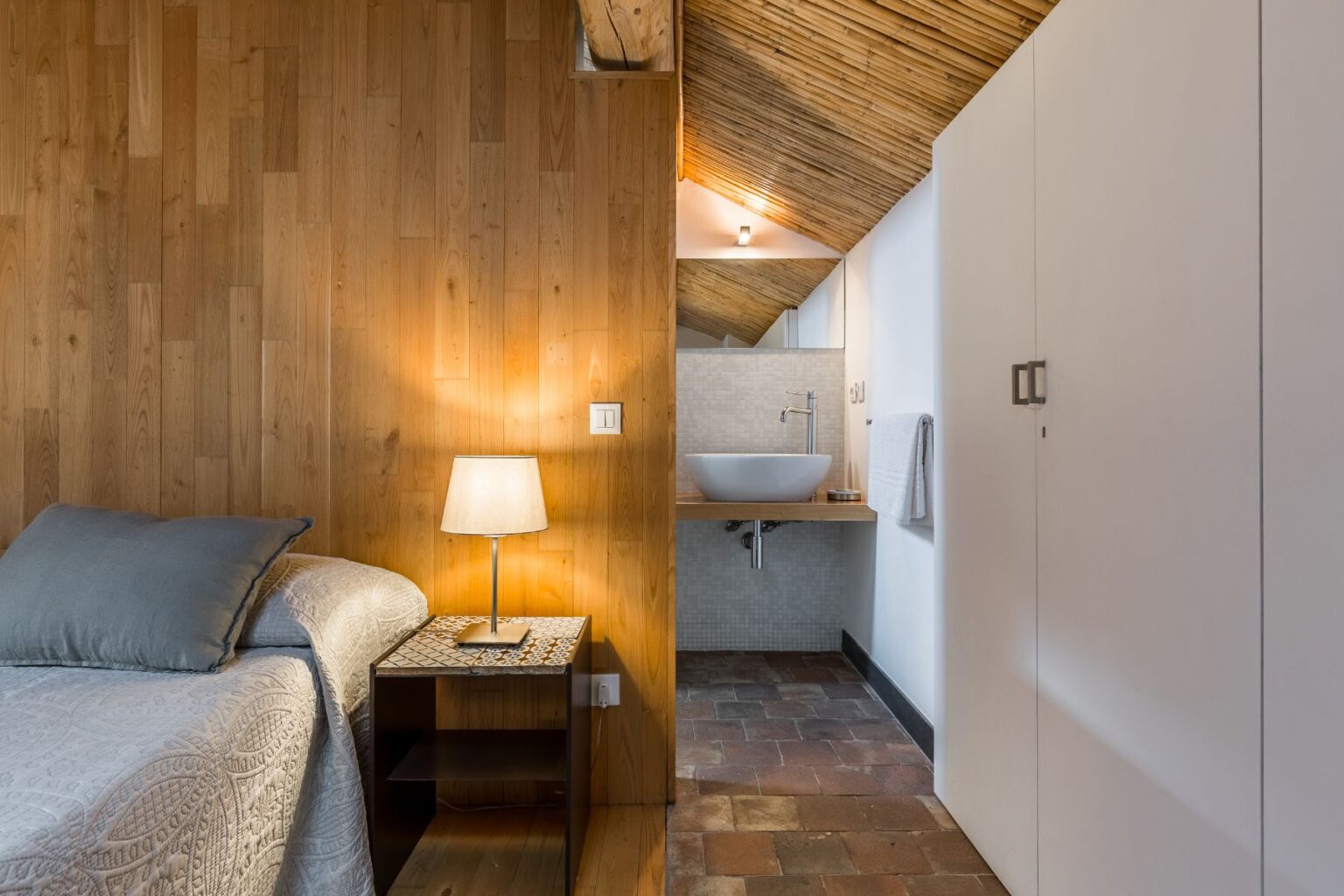 Cozy bedroom with wooden walls, featuring a neatly made bed, a bedside lamp, and a modern bathroom area with a sink and towel rack, showcasing a stylish blend of rustic and contemporary design elements. | Only on SopranoVillas
