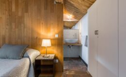 Cozy bedroom with wooden walls, featuring a neatly made bed, a bedside lamp, and a modern bathroom area with a sink and towel rack, showcasing a stylish blend of rustic and contemporary design elements.