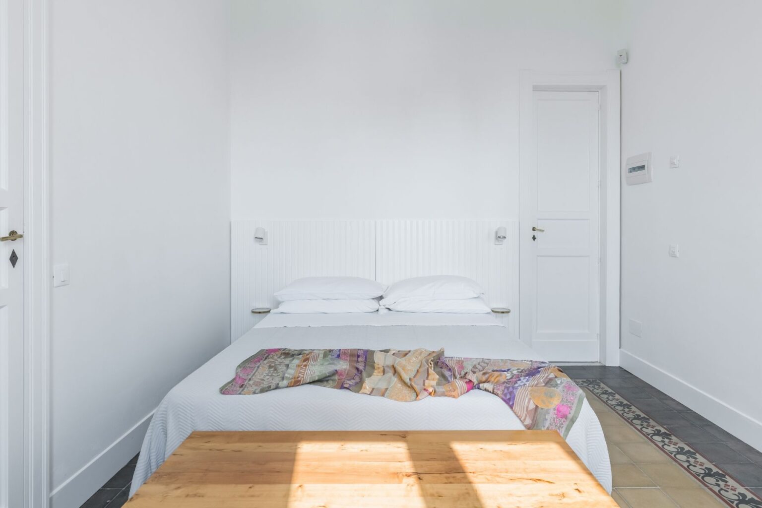 Modern minimalist bedroom featuring a double bed with white linens, a colorful throw blanket, and wooden furniture. The room has light walls, natural light streaming in, and a door leading to another area, creating a serene and inviting atmosphere. Ideal for relaxation and comfort. | Only on SopranoVillas
