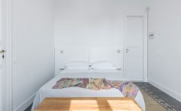 Modern minimalist bedroom featuring a double bed with white linens, a colorful throw blanket, and wooden furniture. The room has light walls, natural light streaming in, and a door leading to another area, creating a serene and inviting atmosphere. Ideal for relaxation and comfort.
