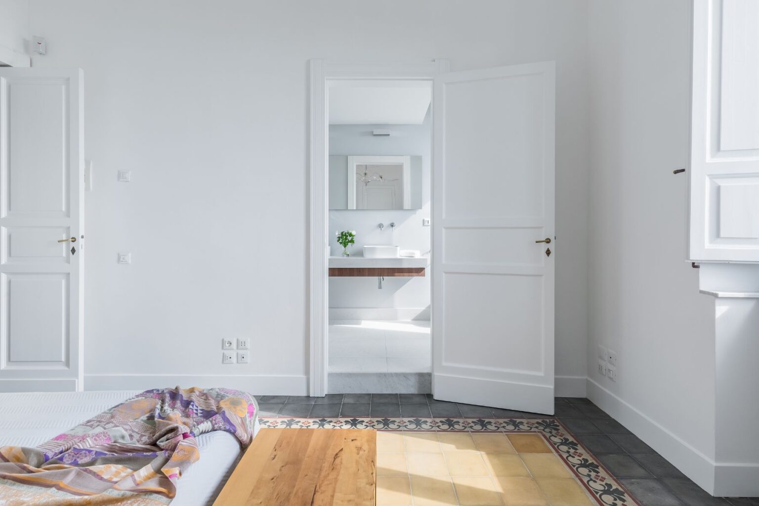 Bright and airy bedroom interior featuring a bed with a colorful throw, wooden bedside table, and an open door leading to a modern bathroom with a sleek vanity and natural light. The room showcases minimalist design with white walls and patterned tile flooring. | Only on SopranoVillas