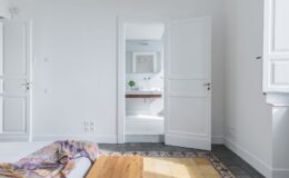 Bright and airy bedroom interior featuring a bed with a colorful throw, wooden bedside table, and an open door leading to a modern bathroom with a sleek vanity and natural light. The room showcases minimalist design with white walls and patterned tile flooring.