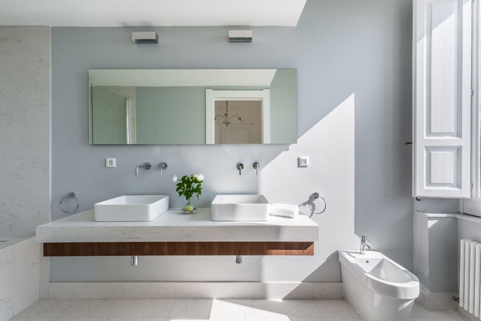 Modern bathroom interior featuring dual vessel sinks on a minimalist countertop, large wall mirror, elegant lighting, and a bidet. Sunlight illuminates the space through a window, highlighting the clean lines and neutral color palette. | Only on SopranoVillas