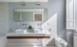 Modern bathroom interior featuring dual vessel sinks on a minimalist countertop, large wall mirror, elegant lighting, and a bidet. Sunlight illuminates the space through a window, highlighting the clean lines and neutral color palette.