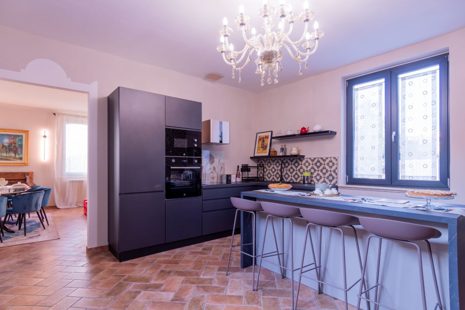 Modern kitchen interior featuring a sleek black refrigerator, built-in oven, and stylish bar stools. The space is illuminated by an elegant chandelier and showcases patterned backsplash tiles, open shelving with kitchenware, and a cozy dining area visible through an archway. | Only on SopranoVillas