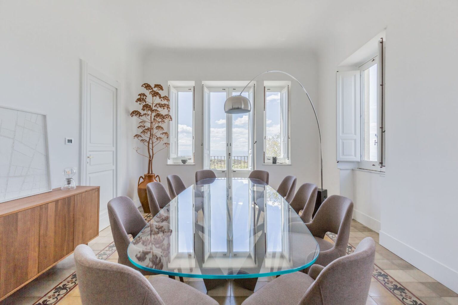 Modern dining room featuring a large glass table surrounded by stylish chairs, with large windows allowing natural light and a decorative potted plant in the corner. The minimalist decor includes a wooden sideboard and an arc floor lamp, creating an inviting atmosphere. | Only on SopranoVillas