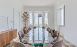 Modern dining room featuring a large glass table surrounded by stylish chairs, with large windows allowing natural light and a decorative potted plant in the corner. The minimalist decor includes a wooden sideboard and an arc floor lamp, creating an inviting atmosphere.