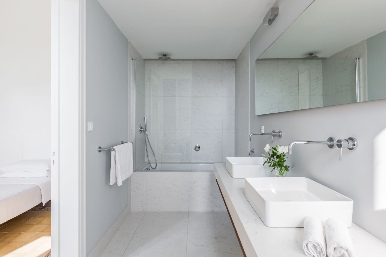 Modern bathroom design featuring a spacious layout with a glass shower, elegant marble walls, dual vessel sinks, and stylish chrome fixtures. The room is well-lit with natural light and includes decorative elements like fresh flowers and neatly arranged towels. | Only on SopranoVillas