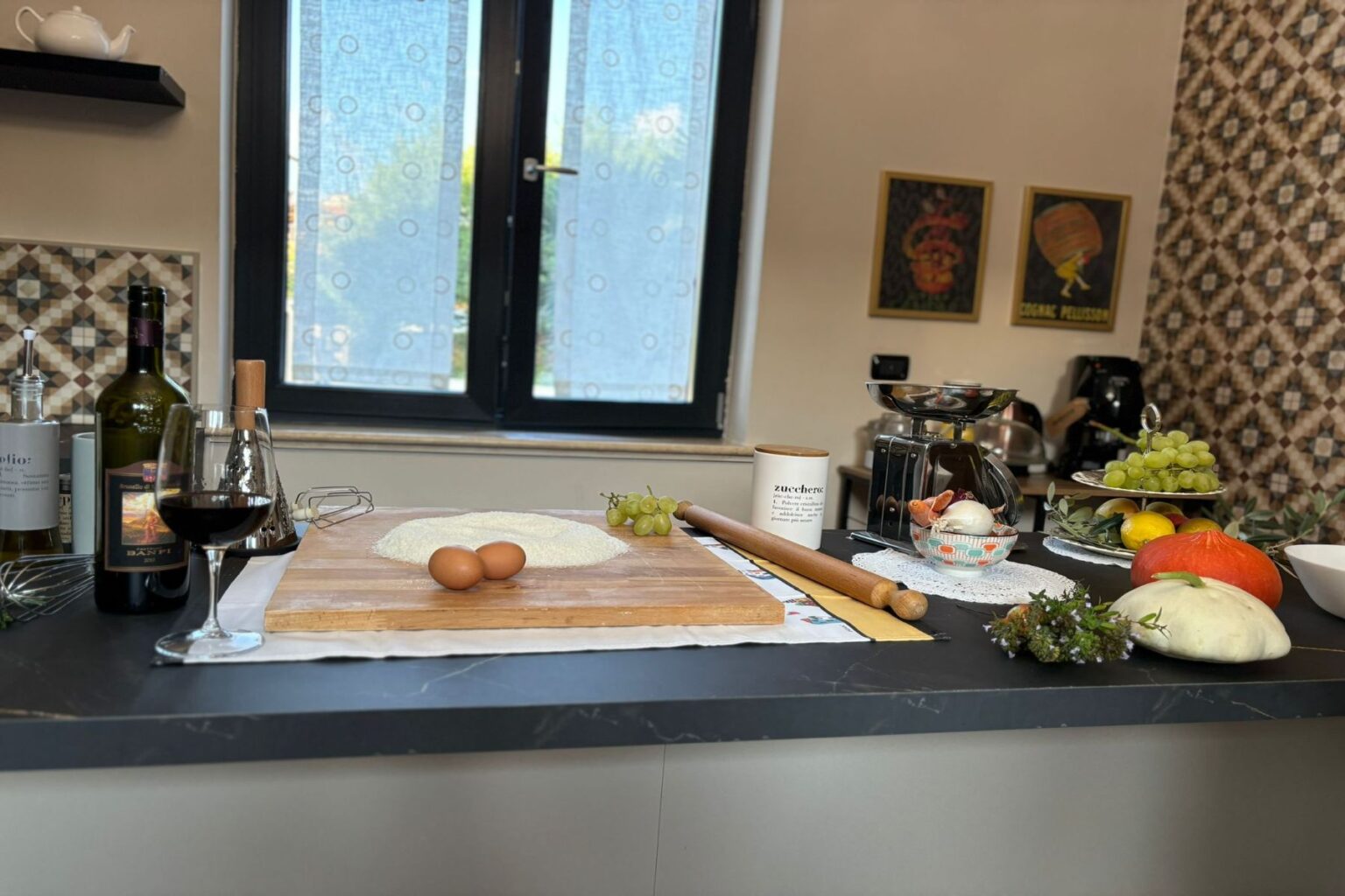 A modern kitchen countertop featuring a variety of cooking ingredients and utensils, including flour, eggs, green grapes, a rolling pin, and a bottle of red wine. The background showcases a window with sheer curtains and decorative artwork on the walls, creating a warm and inviting cooking atmosphere. | Only on SopranoVillas