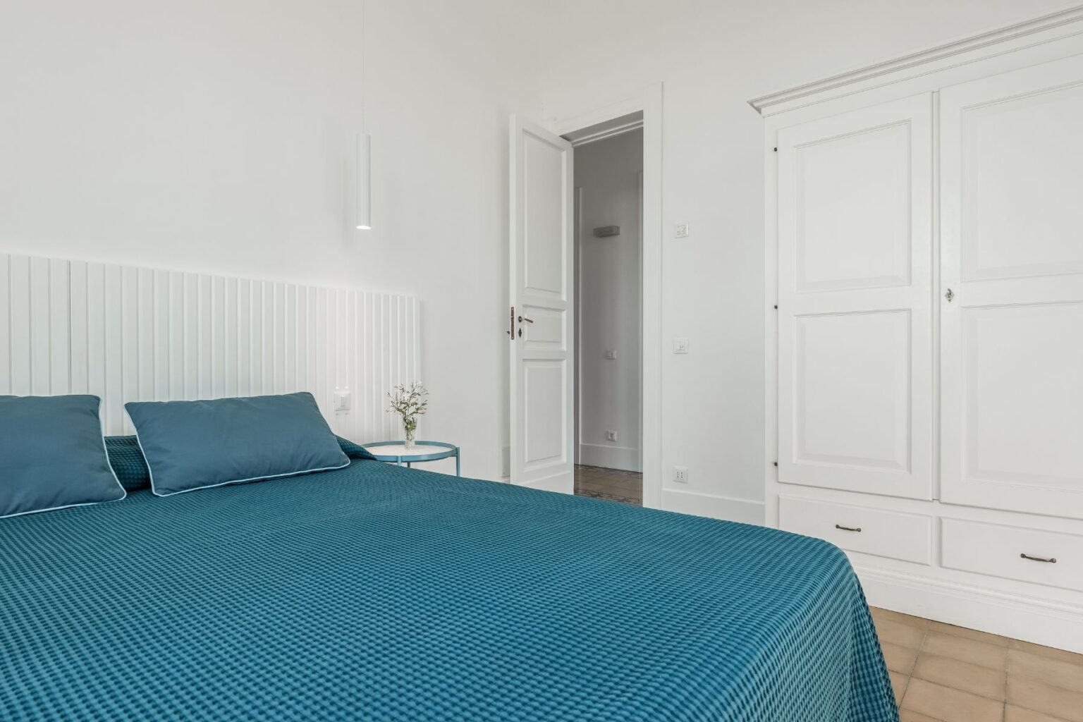 Modern bedroom featuring a teal bedspread and matching pillows, complemented by a white paneled headboard. A minimalist nightstand with a small flower arrangement sits beside the bed, while a white wardrobe and an open door to another room are visible in the background. Natural light brightens the space, enhancing its airy feel. | Only on SopranoVillas