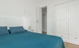 Modern bedroom featuring a teal bedspread and matching pillows, complemented by a white paneled headboard. A minimalist nightstand with a small flower arrangement sits beside the bed, while a white wardrobe and an open door to another room are visible in the background. Natural light brightens the space, enhancing its airy feel.