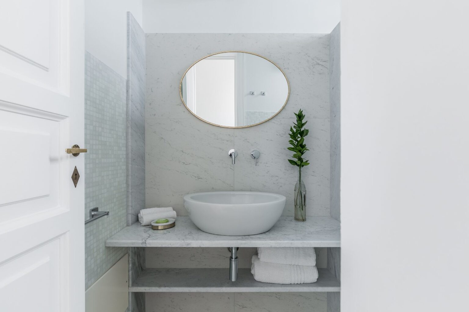 Modern bathroom design featuring a marble sink with an oval mirror, neatly arranged towels, and a decorative plant in a glass vase. The minimalist aesthetic is highlighted by clean lines and a light color palette, creating a serene atmosphere. | Only on SopranoVillas