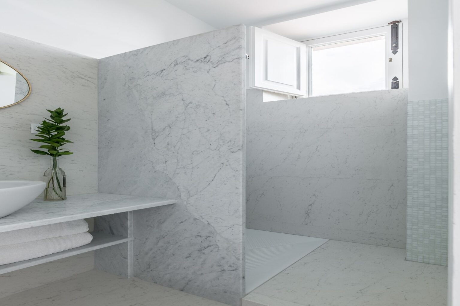 Modern bathroom featuring a sleek marble shower with a glass door, a minimalist sink, and a stylish mirror. Natural light streams in through an open window, enhancing the serene atmosphere. A decorative plant adds a touch of greenery to the elegant design. | Only on SopranoVillas