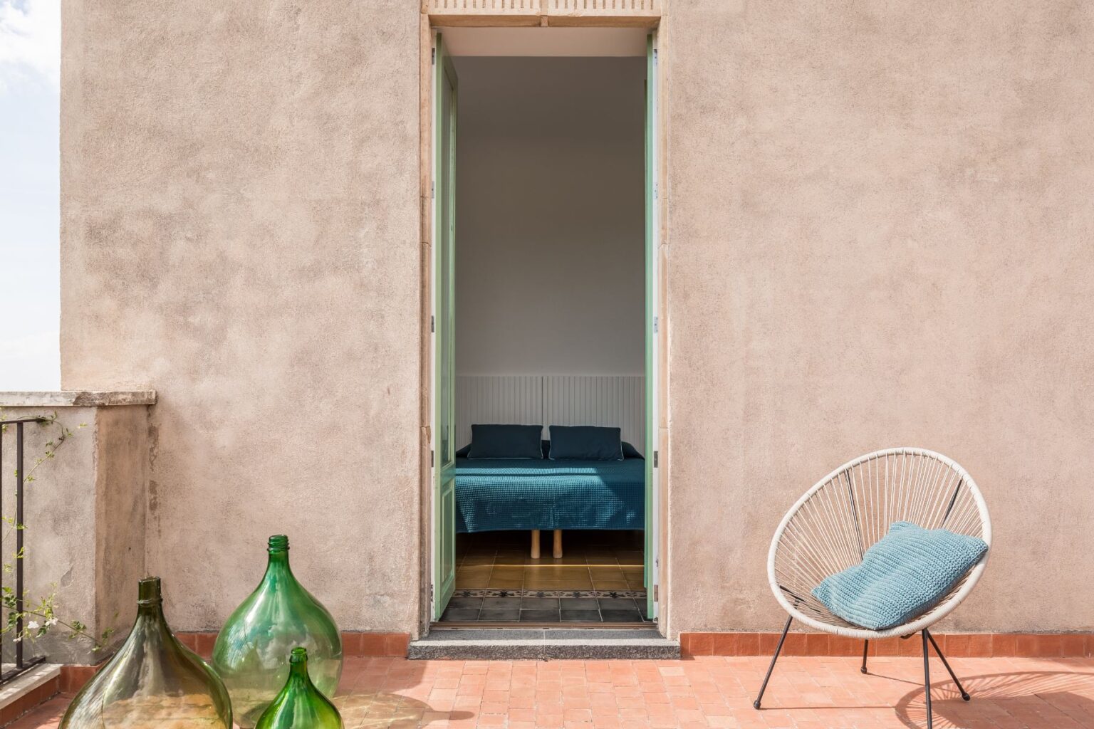 Interior view of a modern bedroom through an open door, featuring a blue bedspread and pillows, with a stylish outdoor seating area and decorative green glass bottles in the foreground. The walls are textured and the floor tiles add a contemporary touch. | Only on SopranoVillas