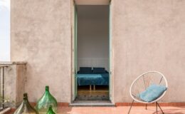 Interior view of a modern bedroom through an open door, featuring a blue bedspread and pillows, with a stylish outdoor seating area and decorative green glass bottles in the foreground. The walls are textured and the floor tiles add a contemporary touch.
