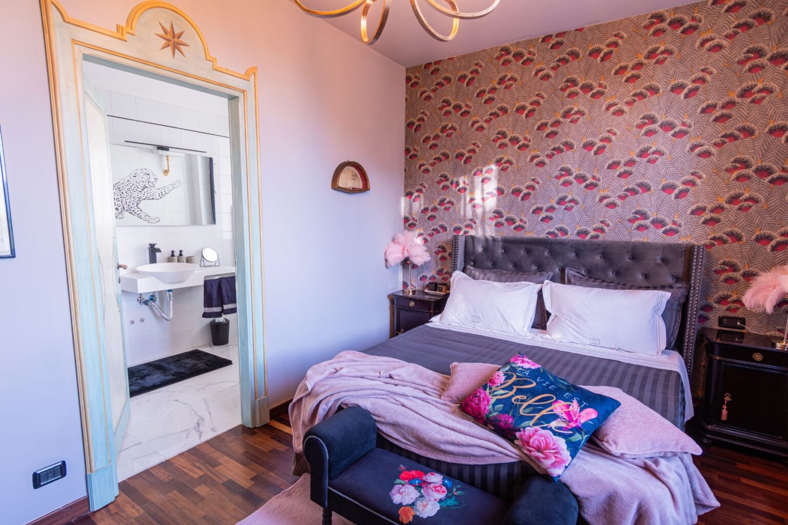 Stylish bedroom featuring a plush bed with decorative pillows, a cozy throw blanket, and a vibrant floral wallpaper. The room includes an elegant chandelier, a small seating area with a dark velvet chair, and a glimpse of a modern bathroom through an ornate doorway. | Only on SopranoVillas