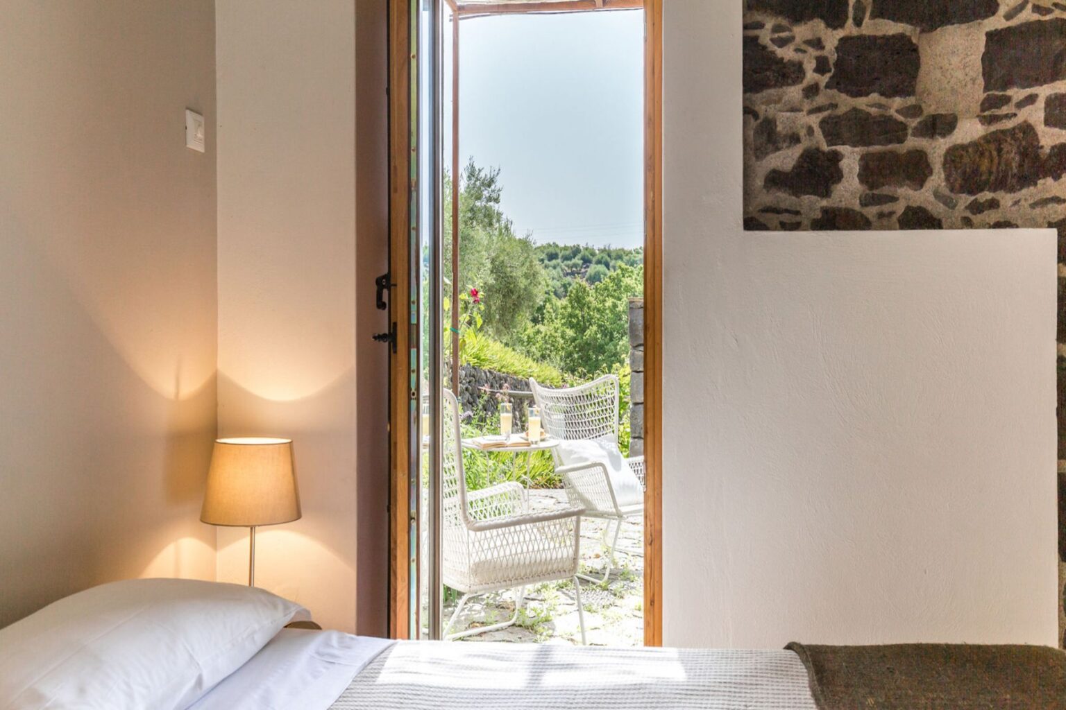 Cozy bedroom with a bedside lamp, featuring an open door leading to a scenic outdoor patio with white chairs and greenery, perfect for relaxation and enjoying nature. | Only on SopranoVillas
