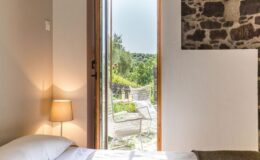 Cozy bedroom with a bedside lamp, featuring an open door leading to a scenic outdoor patio with white chairs and greenery, perfect for relaxation and enjoying nature.