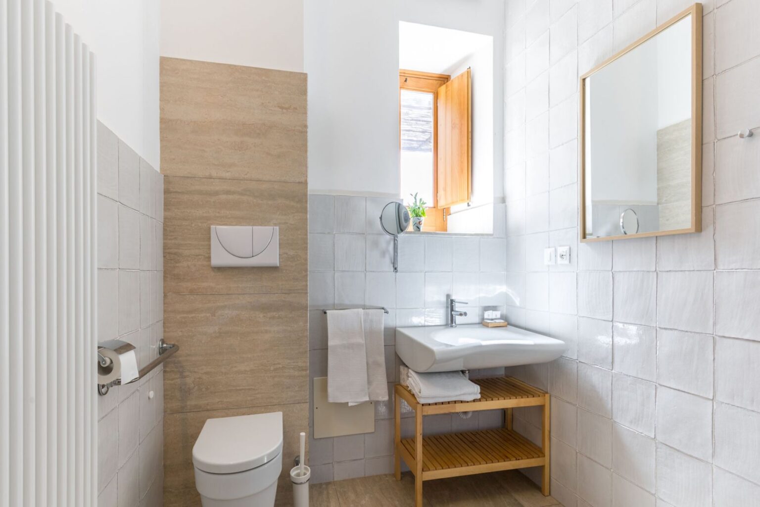 Modern bathroom featuring a wall-mounted toilet, sleek sink with wooden shelving, and natural light from a window. The walls are tiled in a combination of light gray and beige, creating a contemporary aesthetic. A round mirror and towel rack add functionality to the space. | Only on SopranoVillas