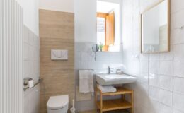 Modern bathroom featuring a wall-mounted toilet, sleek sink with wooden shelving, and natural light from a window. The walls are tiled in a combination of light gray and beige, creating a contemporary aesthetic. A round mirror and towel rack add functionality to the space.