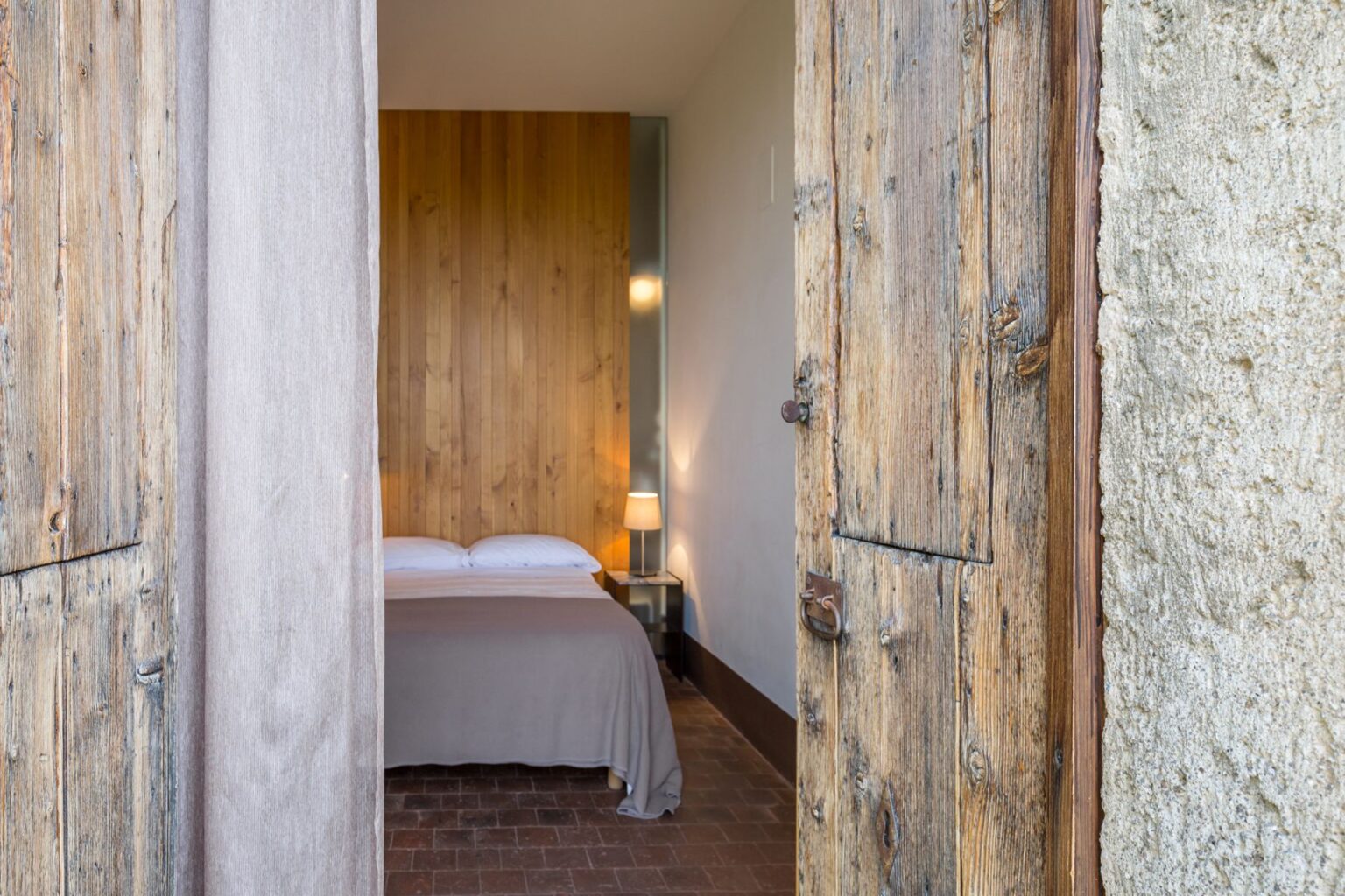 Cozy bedroom interior with a wooden wall and a soft bedspread, viewed through rustic wooden doors. The room features a bedside lamp and warm lighting, creating a welcoming atmosphere. Ideal for showcasing comfortable hotel accommodations or home decor inspiration. | Only on SopranoVillas